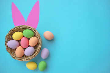 Top view of colorful Easter eggs in basket with pink bunny ears on blue background with copy space ...