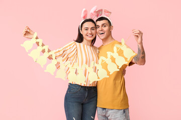 Happy young couple with Easter bunny ears and garland on pink background