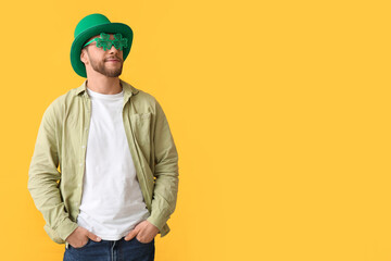 Young man in leprechaun hat and decorative glasses in shape of clover on yellow background. St....