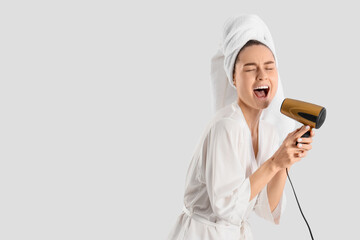 Beautiful woman with towel and hair dryer singing after shower on light background