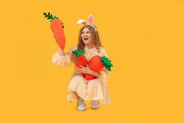 Beautiful young shocked woman in bunny ears with carrot-shaped toys on yellow background. Easter celebration