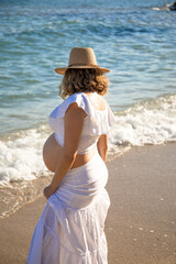 Pregnant woman with sun hat during her walk on the beach relaxing on her summer vacation, happy pregnancy outdoors.