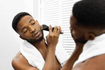 Man grooming beard with trimmer in mirror in bathroom - obrazy, fototapety, plakaty