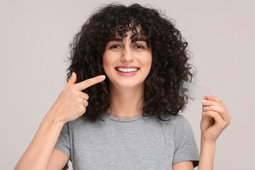 Young woman holding whitening strips and pointing at her teeth on light grey background