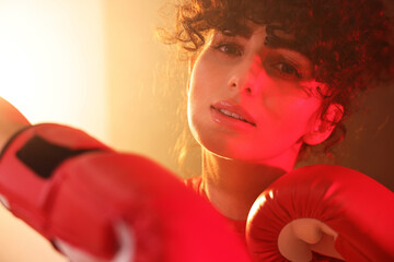 Beautiful young woman with boxing gloves on color background in neon lights and smoke, closeup