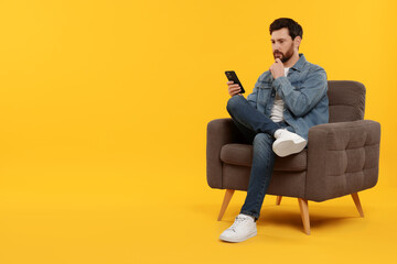 Handsome man with smartphone sitting on armchair against yellow background. Space for text