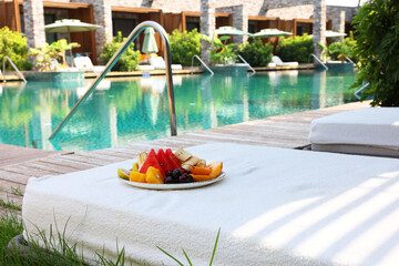 Plate with fresh fruits on sun lounger near outdoor swimming pool. Luxury resort