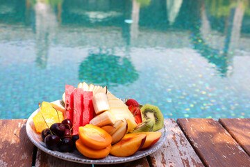 Plate with fresh fruits on wooden deck near outdoor swimming pool, space for text. Luxury resort