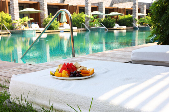 Plate with fresh fruits on sun lounger near outdoor swimming pool. Luxury resort