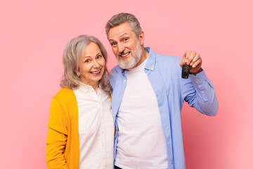 Fototapeta premium Happy pensioners couple excitedly showing automobile key on pink backdrop