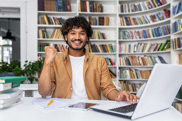 Joyful young man triumphantly clenches fist looking at laptop in library, expressing excitement for his achievement or win.