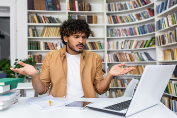 Perplexed young student sitting with a laptop in a library, surrounded by books, looking for assistance.