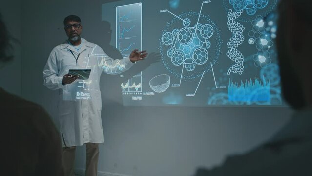 Full footage of middle-aged Asian male clinician with tablet computer standing next to projector screen at meeting, making report about new immunobiological drug, explaining theory to colleagues