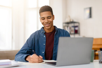 Man smiling while writing notes, laptop open