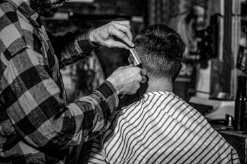 Barber guy gives a haircut to a bearded man sitting in a chair in a barbershop. Barber preparing hair clipper for bearded man, barbershop background. Barber works with a beard clipper - obrazy, fototapety, plakaty
