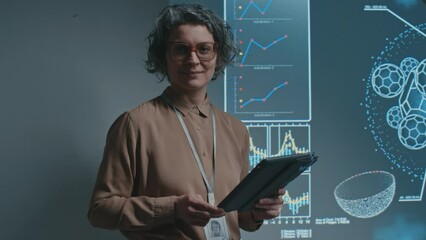 Medium portrait shot of Caucasian female scientist or professor with tablet computer and headset microphone standing by projector screen, giving lecture on biochemistry, turning to camera and smiling