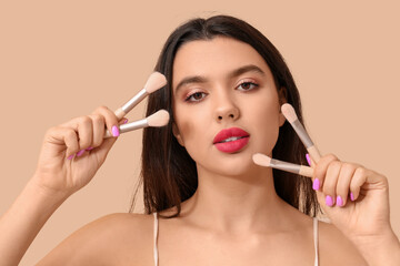 Beautiful young woman with makeup brushes on beige background - obrazy, fototapety, plakaty