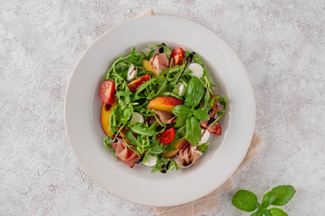 Salad with peaches, arugula, prosciutto, mozzarella cheese and cherry tomatoes with balsamic vinegar in a ceramic plate on a concrete background. Selective focus.