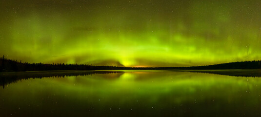 Spectacular aurora in many shades of green and hints of orange and red reflect in a large calm lake. The shoreline of the lake has silhouettes of a dark forest. Stars can be seen through the aurora.