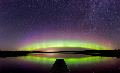 Aurora and stars are reflecting in a northern lake that has a long dock in the foreground.  The...