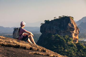 Man with backpack sitting on rock and looking at landscape. Scenery with Sigiriya rock in Sri Lanka. . - obrazy, fototapety, plakaty