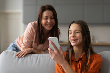 Pretty teenage girl sharing mobile phone to mother, talking on online video call, smiling, watching content on Internet