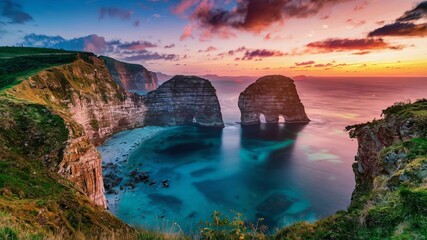 Beautiful beach with high cliffs and a blue sea at sunset. The sky is orange and pink. The cliffs are brown and grey, and there are two large rocks in the sea, which appear grey.