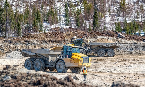 Two dump trucks transporting sand to a construction site, with a surveyor carrying out a survey