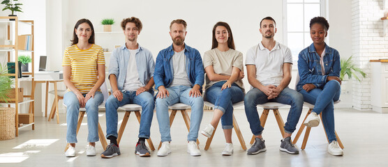 Group of diverse people sitting on chairs in row. Front view portrait of male and female candidates in casual denim clothes waiting in line in office before job interview