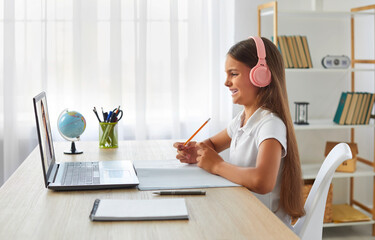 Happy preteen schoolgirl in headphones studying online at virtual class with teacher tutor. Side view of smiling Caucasian child girl sitting at home in front of laptop and listening to teacher. - obrazy, fototapety, plakaty
