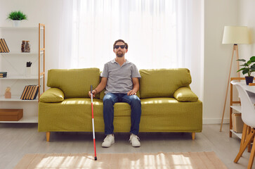 Portrait of a young blind man with stick cane sitting on sofa in the living room at home. Male...