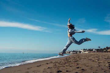 A tanned young woman jumps in a blue suit, a brunette in a tracksuit, an active girl jumps cheerfully on the sea coast. Athletic girl in a tracksuit jumping on the sand - Powered by Adobe