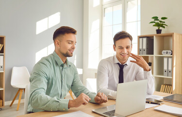 Obraz premium Business men at work in office. Two young confident employees sitting at the desk on meeting looking at laptop monitor screen discussing new projects or startups, analyzing company.