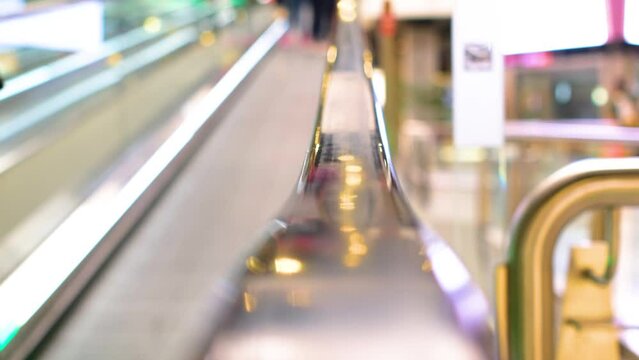 Centered shot of the detail of the handrail of an escalator in a shopping mall. In the background pass out of focus people anonymously shopping.