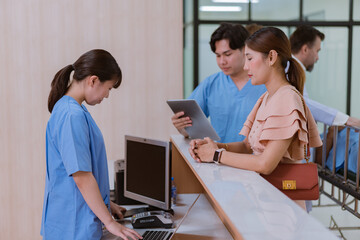 Asian female administrator talks with patient. Adult woman stands near reception desk in clinic lobby area, asks information, makes appointment with doctor. Medical staff work in modern hospital.