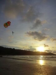 The tranquil ocean at sunset, framed by a dark mountain silhouette, gentle light peeking through the clouds, and a paragliding adventure in the distance.