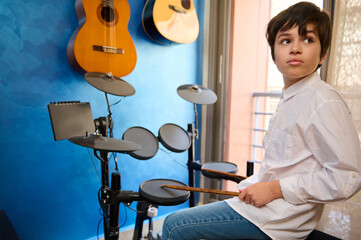 Teenager boy drummer musician sitting at drum set in his retro music studio at home, holding...
