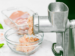 Manual retro meat grinder with minced meat in a glass bowl with boneless chicken thighs and basil leaves