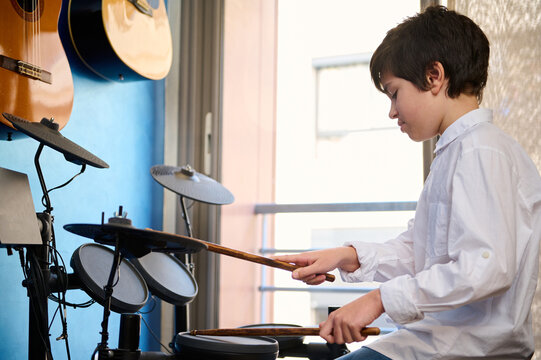 Side portrait of a Caucasian handsome teenage boy musician drummer banging on drums at home music studio. Adolescent boy beating on cymbals with drumsticks, sitting at modern drum equipment