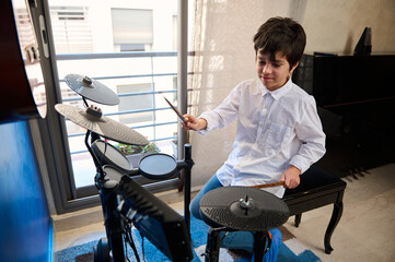 Teenager boy playing drums at home music studio, holding drumsticks and banging drums, beating on...