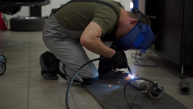 Auto mechanic welds exhaust pipe in workshop. Welding sparks fly from flex pipe as repairman fixes vehicles exhaust system. Technician in safety gear works on car catalyst rejuvenation.