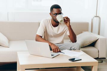 Smiling African American Freelancer Working on Laptop in Modern Home Office while Sitting on Sofa This image captures a young African American guy, dressed casually, sitting comfortably on a couch in