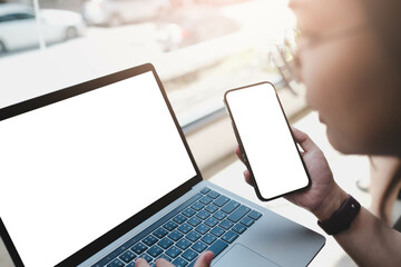 Close-up image of a woman sitting in the cafe and using her smartphone and laptop. smartphone and laptop white blank screen mockup for display your graphic banner. - obrazy, fototapety, plakaty
