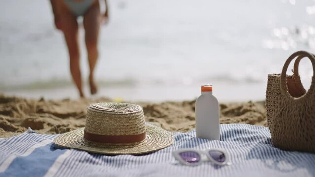 Sandy beach adorned with summer essentials UV protection sun cream, woven straw hat, trendy sunglasses, elegant straw bag, blue towel. Ocean backdrop with person walking towards water. Slow motion.