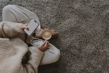Cozy morning routine. Top view of woman wearing pastel creamy clothes sitting on fluffy grey carpet with cup of coffee and using mobile phone with blank screen. Social media mockup with copy space - 772367273