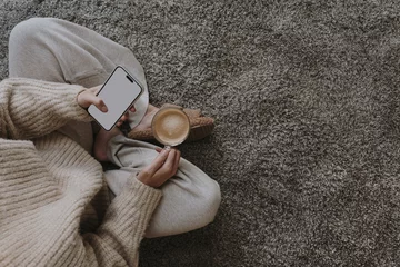 Foto op Plexiglas Cozy morning routine. Top view of woman wearing pastel creamy clothes sitting on fluffy grey carpet with cup of coffee and using mobile phone with blank screen. Social media mockup with copy space © Floral Deco
