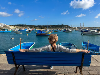 Woman sitting on bench and looking at boats in Marsaxlokk harbor, Malta