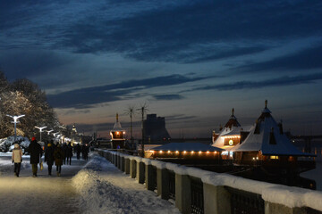 A winter and night city with people, benches, houses, buildings, white snow, bare trees, lanterns are located along the Dnipro River.