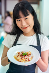 An Asian woman proudly wears an apron with a beautifully arranged plate of food ready to be served.