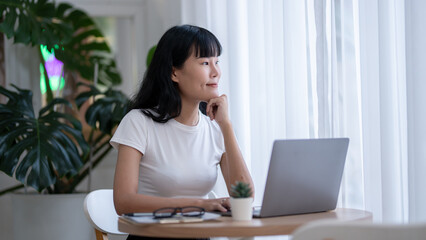 Asian woman sitting contemplating holding a coffee cup while looking away from her laptop. Looking...